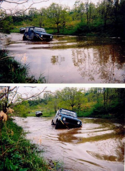 1978 Ford Bronco - Two off-road vehicles navigate through a muddy waterway surrounded by lush green trees.