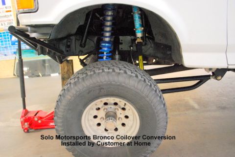 1981 Ford Bronco - A white Ford Bronco's front suspension is shown with a large tire and a red hydraulic jack underneath. The image highlights a coilover conv…