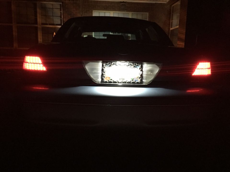 Rear view of a dark sedan at night, with red taillights and license plate light on.