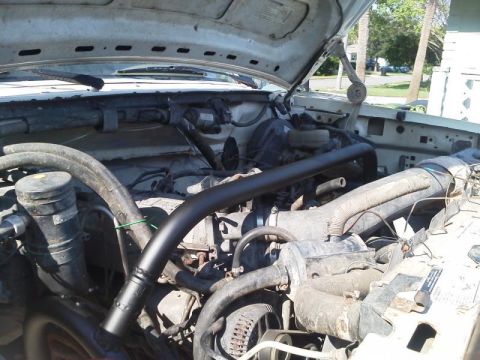 1981 Ford Bronco - The open hood of a white Ford truck reveals a dusty engine bay with various hoses, pipes, and the air intake system visible. A black curved…