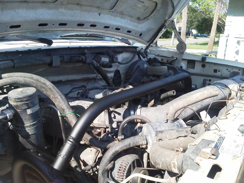 Engine bay of a white Ford truck with open hood, showing hoses and black pipe.