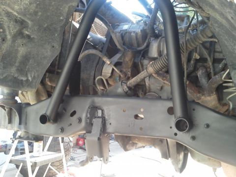 1981 Ford Bronco - The underside of a vehicle frame with newly painted black support bars and brackets. The engine and various hoses are visible in the backgr…