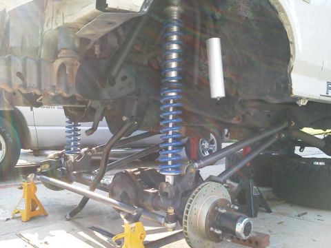 1981 Ford Bronco - The underside of a lifted 1981 Ford Bronco is shown, with the front suspension components exposed. Blue coilover shocks and heavy-duty link…