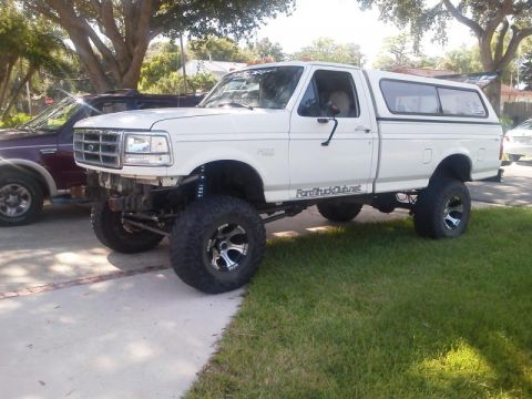 1981 Ford Bronco - A lifted white Ford F-250 pickup truck with a camper shell is parked on a paved driveway next to a dark red Ford Bronco. The front bumper o…