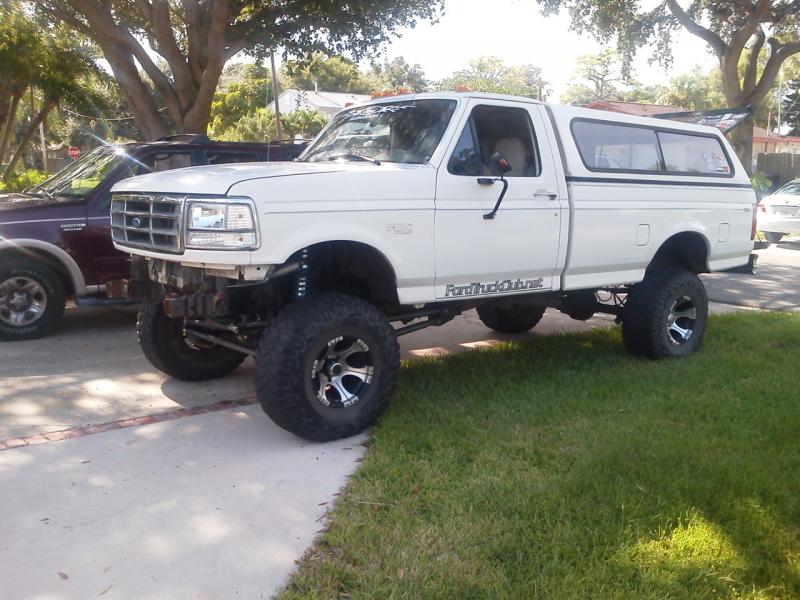 Lifted white Ford F-250 pickup truck with camper shell, front bumper removed, parked on driveway.
