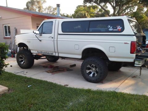 1981 Ford Bronco - A lifted, white Ford pickup truck with a white camper shell is parked on a concrete driveway in front of a house. The truck has large off-r…