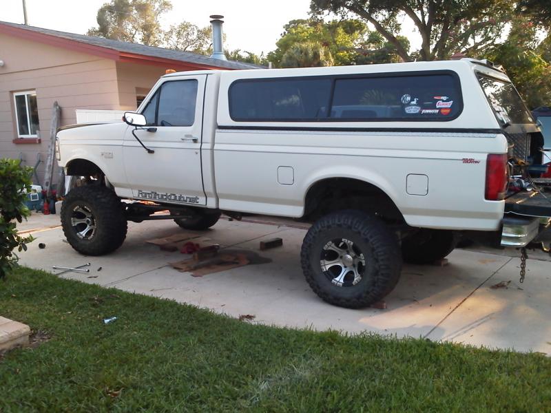 Lifted white Ford pickup truck with camper shell and off-road tires parked on driveway.