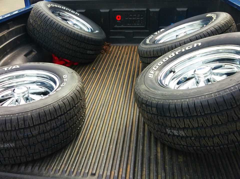 Four BFGoodrich radial tires with chrome rims in the bed of a blue pickup truck.