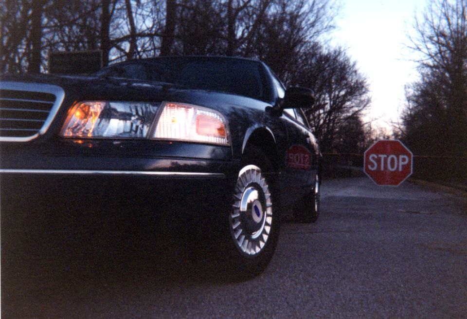 Low-angle front-left of dark sedan with chrome wheel near a STOP sign at dusk