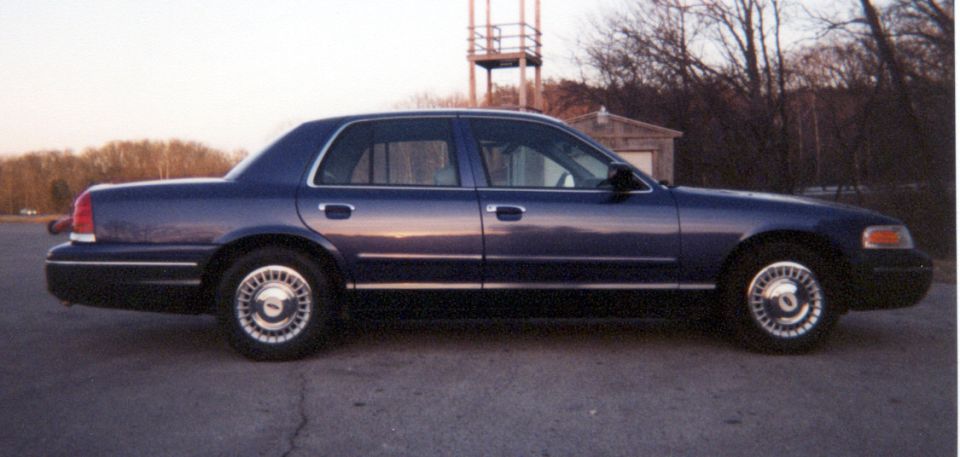 Side view of dark blue Ford Crown Victoria sedan parked on pavement with bare trees behind