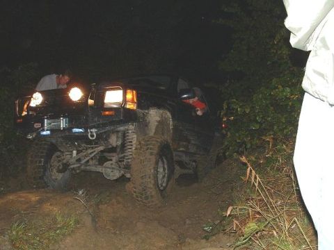 1978 Ford Bronco - A black off-road SUV navigates a challenging muddy trail in the dark, with auxiliary lights illuminating the path.