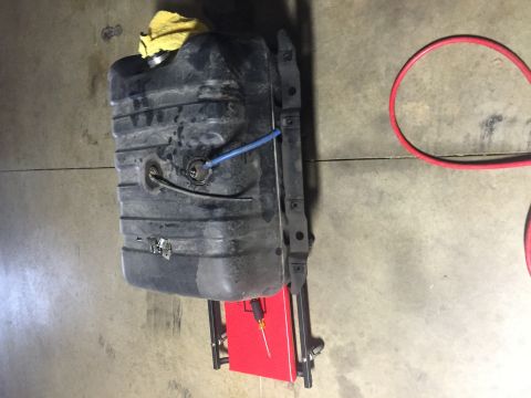 1982 Ford Bronco - A black plastic fuel tank, possibly from a 1982 Ford Bronco, is mounted on a red stand with a blue hose attached. A yellow cloth is visible…