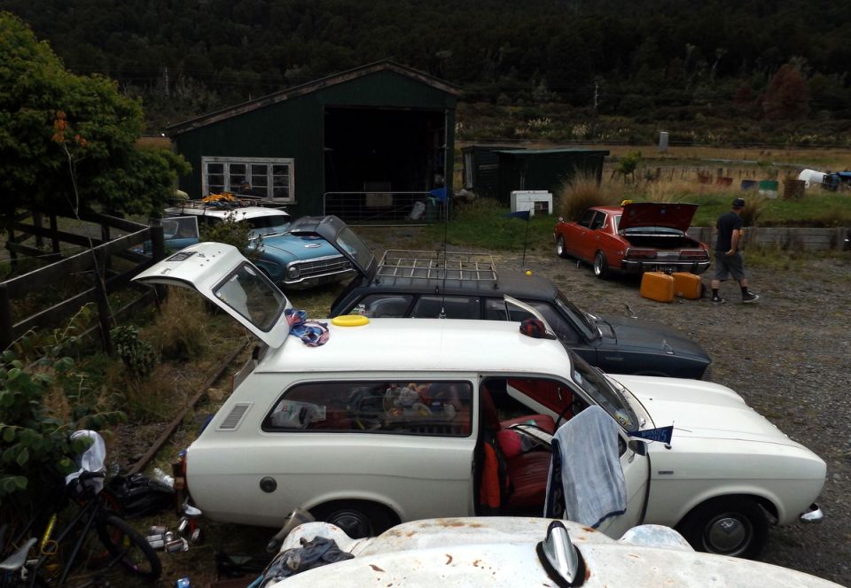 Several vintage cars parked in front of a barn, including a white station wagon with open tailgate.