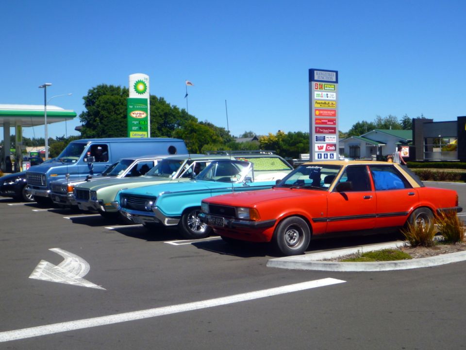 Row of vintage cars including a blue van, green sedan, turquoise sedan, and red sedan with tan roof.