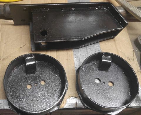 1991 Ford Bronco - Two black metal spring perch cups with mounting tabs and holes are shown in the foreground, with a black metal bracket assembly in the back…