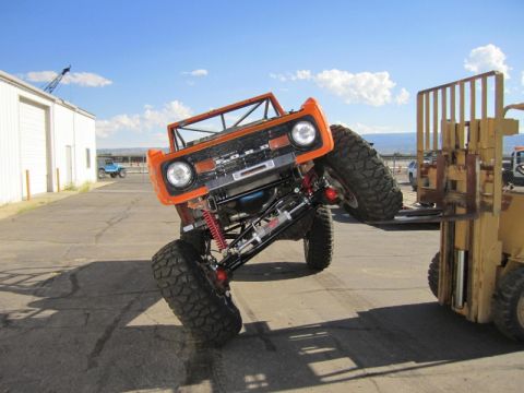 1981 Ford Bronco - An orange Ford Bronco is tilted on its side, with its front tires resting on the forks of a yellow forklift. The vehicle is in an outdoor p…