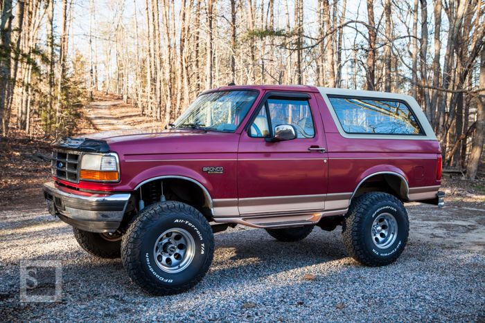 Maroon 1990s Ford Bronco with tan top and off-road tires parked in a wooded area.
