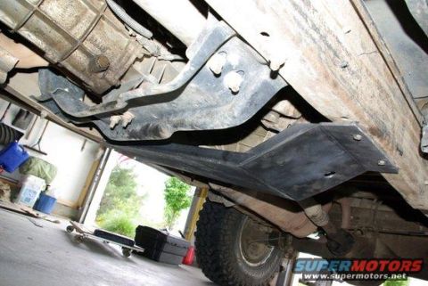 1981 Ford Bronco - The underside of a vehicle shows a black metal skid plate bolted to the frame, protecting the drivetrain components. A portion of the rear…