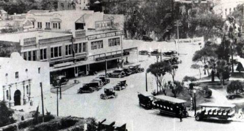 1909 Ford Model T - A bustling street scene captures multiple vintage automobiles navigating a town square with historic architecture and lush trees.