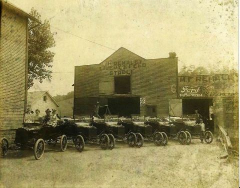 1909 Ford Model T - A row of early automobiles are parked outside a livery and Ford dealership, with people gathered around some of the vehicles.