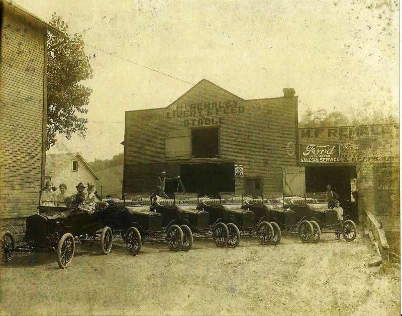 Several early cars parked in front of a building labeled 'J.H. REMALEY LIVERY & FEED STABLE' and 'M.F. REMALEY Ford SALES & SERVICE'