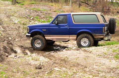 1996 Ford Bronco - Blue 1996 Ford Bronco navigates a muddy off-road trail with a spare tire mounted on the rear.