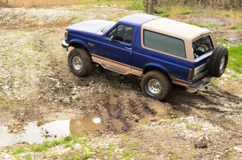 1996 Ford Bronco - This blue Ford Bronco is navigating a muddy trail with puddles and rough terrain.