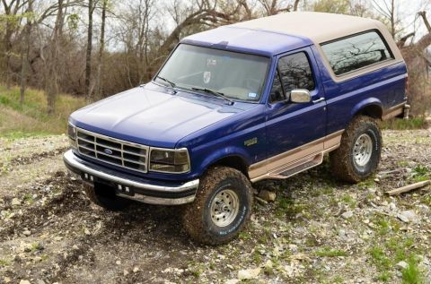 1996 Ford Bronco - This blue Ford Bronco is parked on a dirt trail surrounded by trees and brush.