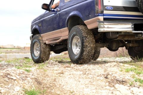1996 Ford Bronco - A blue 1996 Ford Bronco is shown from the rear 3/4 angle, equipped with large off-road tires and driving on a dirt path.