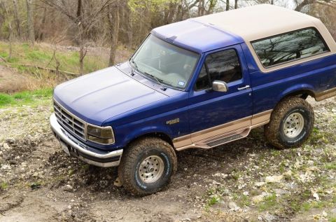 1996 Ford Bronco - A blue Ford Bronco is shown on a muddy, off-road trail with trees in the background.