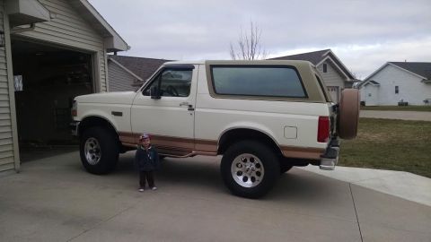 1994 Ford Bronco - A white and tan 1994 Ford Bronco is parked on a concrete driveway next to an open garage. A young child stands in front of the driver's sid…