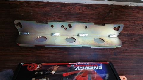 1995 Ford Mustang - A yellow-zinc plated metal bracket with multiple holes and welds sits on a dark wood surface. Below it is a partial view of a black box wit…