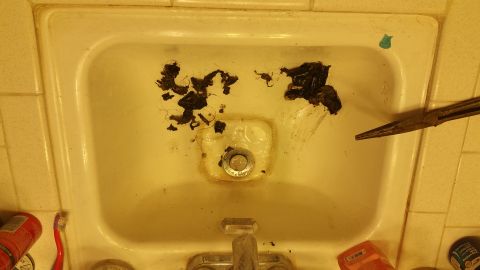 1995 Ford Mustang - A clogged sink drain is being cleared with a pair of needle-nose pliers. The sink basin contains a significant amount of hair and debris.