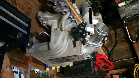 1995 Ford Mustang - A silver aluminum intake manifold with a black throttle body is mounted on a wooden surface, likely in a garage or workshop setting. The ma…