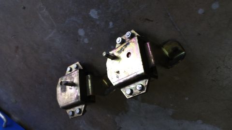 1995 Ford Mustang - Two gold-colored metal engine mounts with bolts are shown on a dark, textured surface. One mount is positioned to the left and slightly for…