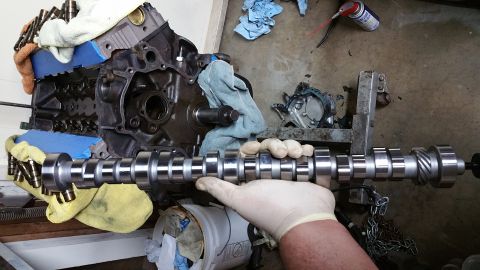 1995 Ford Mustang - A person wearing white gloves holds a camshaft in front of an engine block. The engine block is partially covered with blue tape and yellow…