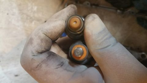 1995 Ford Mustang - A gloved hand holds two fuel injectors, showing their tops with orange seals and metal nozzles.