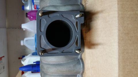 1995 Ford Mustang - A close-up view of a dark gray intake manifold with a black gasket and four silver bolts. The manifold is positioned horizontally, with a c…