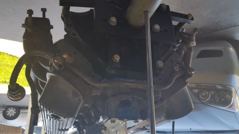 1995 Ford Mustang - The underside of a vehicle's engine and exhaust system is shown, mounted with various bolts and brackets. A silver SUV is visible in the ba…