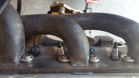 1995 Ford Mustang - Close-up view of a V8 engine's exhaust manifold and spark plugs. The exhaust manifold is made of dark metal with visible welds, and several…