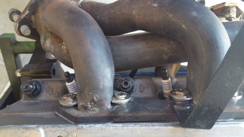 1995 Ford Mustang - A close-up view of a V-shaped engine exhaust manifold with four spark plugs installed on the engine block. The manifold is made of dark met…