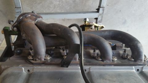 1995 Ford Mustang - A close-up view of a V-shaped exhaust manifold with four exhaust pipes connected to spark plugs on an engine. The manifold is dark metal an…