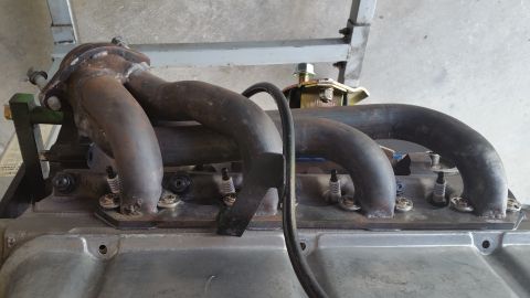 1995 Ford Mustang - A close-up view of an engine's exhaust manifold and spark plugs, attached to the engine block. The exhaust manifold is made of dark metal p…