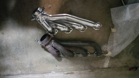 1995 Ford Mustang - Two exhaust manifolds are laid out on a concrete floor. The top manifold is shiny and new, while the bottom one appears older and rusted.