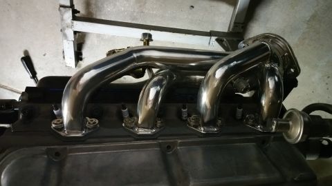1995 Ford Mustang - A polished chrome exhaust manifold is installed on a dark-colored engine block, with spark plugs visible below the manifold's flanges. The…