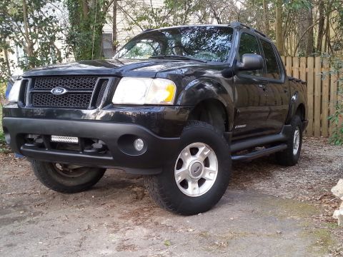2001 Ford Explorer - A black Ford Explorer Sport Trac is parked on a gravel and dirt surface, with a wooden fence and trees in the background. The truck feature…