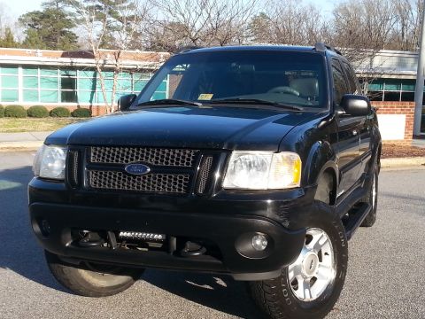 2001 Ford Explorer - A black 2001 Ford Explorer Sport Trac is parked in front of a brick building with large windows. The front of the SUV is visible, featuring…