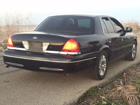2008 Ford Crown Victoria - The rear driver's side of a black Ford Crown Victoria sedan is shown parked on the side of a road. The car has chrome wheels and a chrome t…