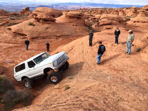 1979 Ford Bronco - A white 1979 Ford Bronco with off-road modifications is navigating a rocky, desert terrain. Several people are standing around observing th…