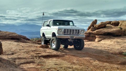 1979 Ford Bronco - A lifted white 1979 Ford Bronco is parked on a rocky, desert landscape with mountains in the background under a cloudy sky. The vehicle has…